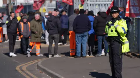PA Media A police officer stands in the foreground on a pavement, looking in our direction. Behind her is a number of people on a picket line in front of the large metal gates of a waste facility.