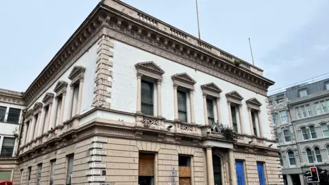 A large square building in Belfast. The building is white and light brown. The windows are boarded up, some are brown and some blue. In the background there are other large buildings, they are grey. In the corner is a traffic light on red. 