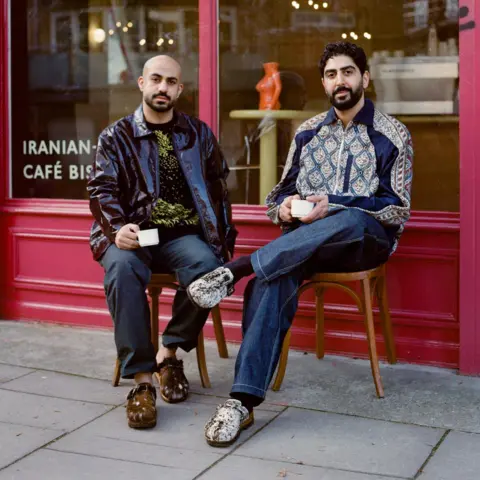 Shams Al Fekaiki Two men sit on chairs holding coffee cups outside a red-painted cafe 