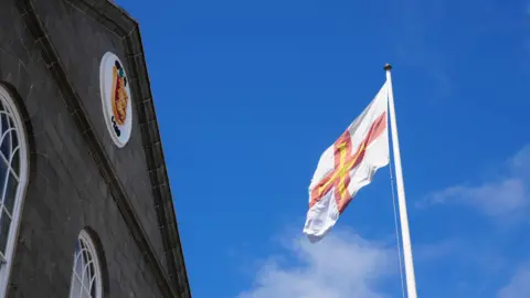 The outside of the States Chamber Building. The top of the building is shown alongside a Guernsey flag on a flag pole. In the background is a clear blue sky.