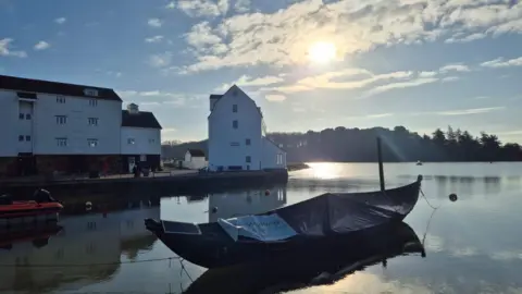 Vikki Irwin/BBC The River Deben on a sunny day in Woodbridge. A small boat rests on the water with white buildings and homes seen behind it. 