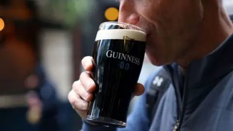 Reuters A man in a blue jacket drinks a Guinness 0.0 zero alcohol beer in a branded glass. You can only see the bottom half of his face