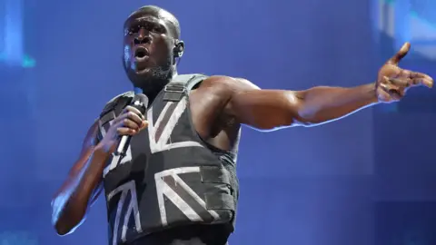 Stormzy performing on stage wearing Banksy's bullet proof vest with a spray painted Union Jack flag on it. He is lit up by a blue hue coming from behind him. He is holding the microphone up to his mouth in his right hand and has his right arm outstretched to pump up the crowd.