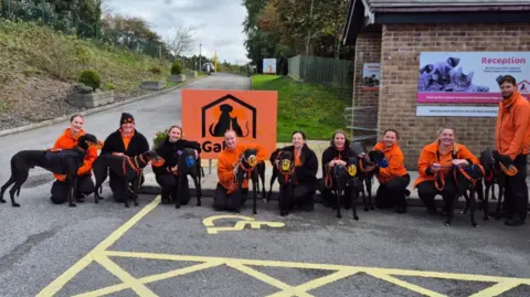 A group of people standing and kneeling outside a brick building with a sign that reads “Reception.” They are all wearing matching orange and black uniforms and are holding several black greyhounds on leads. In the centre, there is a large orange sign featuring a silhouette of a dog and cat inside a house shape. There is also a yellow-painted disabled parking symbol on the ground in front of the group. 