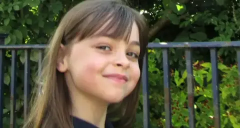 Saffie Roussos standing in front of greenery with a cheeky smile on her face. She has long brown hair with a short fringe