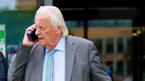 PA Media Michael Mansfield with white hair wearing a grey suit and talking on the phone