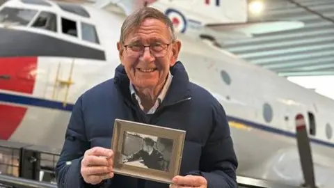 A man wearing a blue coat and shirt and glasses and smiling at the camera. He is holding a black and white photo of himself as a young man wearing an RAF uniform. 