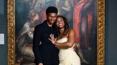 A man and a woman smile holding each other in front of a large oil painting