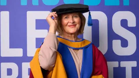 LEEDS BECKETT UNIVERSITY Anne poses for the camera, wearing a university gown and cap. She has a blonde-coloured bob hair and has blue eyes. She stands in front of a purple-painted wall which reads 'Leeds Beckett'.