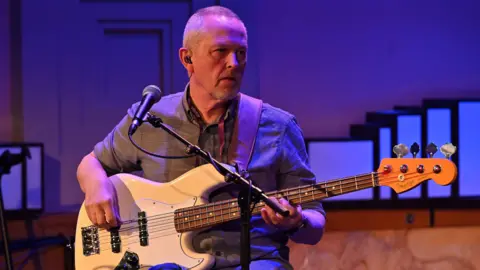 Mark Allan/BBC Horace Panter playing guitar on stage. He has receding grey hair, white stubble and the guitar is cream with a white embossed part. There is a microphone on a stand in front of him.