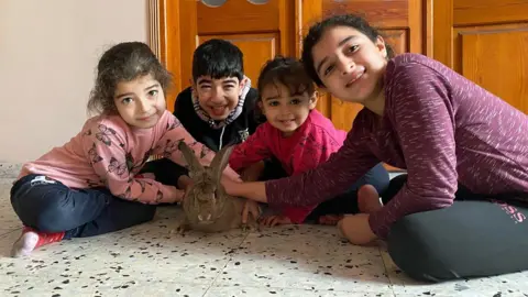 Asmaa Tayeh Four children - three girls and a boy - sitting next to each other crossed legged, all leaning forward and smiling, petting a plump rabbit.