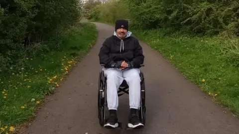 Family A man is sitting in a wheelchair on a tarmac path. Either side of the path is grass he is wearing a black beanie hat and a black zipped up jacket and has a grey tracksuit on.