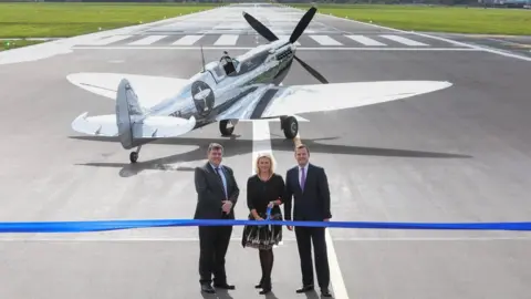 Southampton Airport Three people stand in front of a runway about to cut a blue ceremonial ribbon