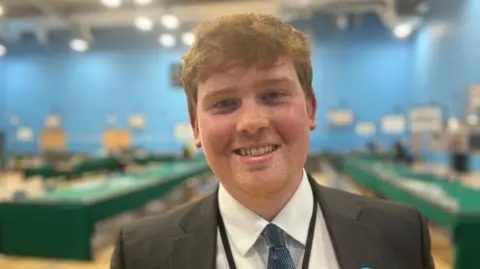 A man with medium-length light brown hair, smiling at the camera and wearing a dark jacket, a white shirt, a blue tie and a lanyard. He is at an election count and tables covered in green cloths are behind him.