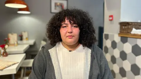 Hadil has dark curly shoulder length hair and wears a grey hoodie over a white ribbed top. She is in a cafe, with small white tables in a row on the left and a hatch which is decorated with hexagonal tiles below, and a woven basket on the hatch shelf.