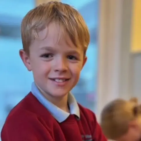 Family handout A boy smiling at the camera. He has blonde, short hair, with a fringe, and is wearing a red school jumper with a light-blue collar poking over the top. The background is blurred, but you can see a window.