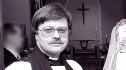 AFP A vicar dressed in vestments and wearing 1980s-style glasses looks into the camera against the backdrop of a church altar and a cross above it. The image is monochrome.