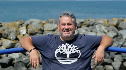 Colin Hattersley A man with greying black hair and a beard in a blue patterned T-Shirt leans with his arms on a blue metal railing in front of a rocky seafront with sea behind