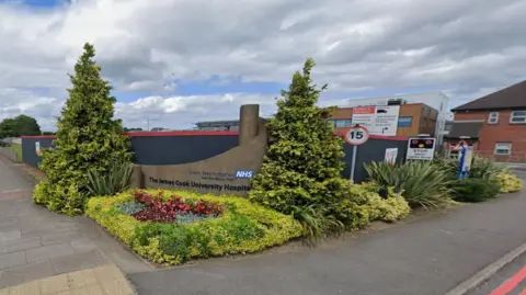 The entrance to the James Cook University Hospital. A wall displaying the hospital's name is surrounded by flowers and shrubs.