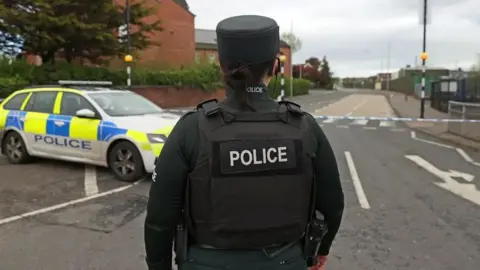 A police officer standing with their back towards the camera. They are wearing green and have their hair tied up. They also have a hat on. There is a police car and cordon on the street in front of them. There is a zebra crossing behind the cordon. Red brick buildings are in the distance, behind a tree and hedge. 