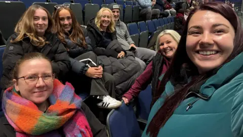 Laura McCloskey A group of people wearing coats and sitting in a hockey arena all smiling to the camera.