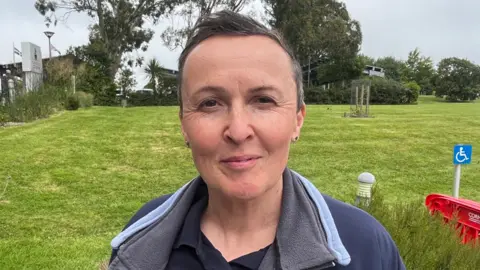A picture of a lady with short hair and smiling at the camera. There is a large grass open space behind with signs and trees in the distance.