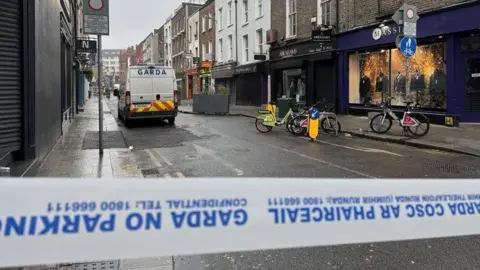 A street is cordoned off with Garda tape in Dublin city centre. A Garda van is in the street as are a number of shops and bikes.