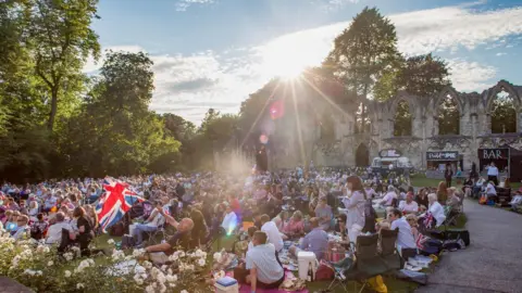 Hundred of people sat on the grass on camping chairs, some are sat on the floor at the York Museum Gardens for the York Proms with the sun coming over the ruins. 