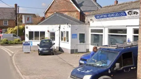 Two cars parked outside a white building with a blue sign next to a road. 