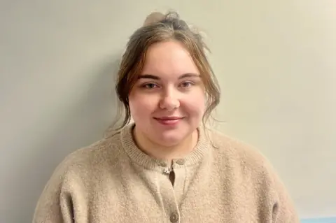 Alana More Alana More, a young woman with dark blonde hair tied up. She is wearing an oatmeal-coloured cardigan