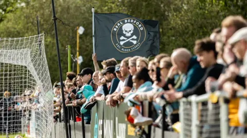 A line of football fans standing behind hoardings with a flag flying in the background, with the logo of the team "Real Bedford".