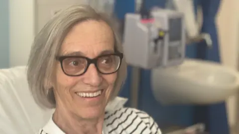 Chris smiles for the camera. She has grey bobbed hair and wears dark-framed glasses.