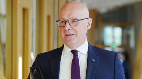 PA Media John Swinney, who is bald with glasses, walks while holding a folder under his arm. He is wearing a business suit 