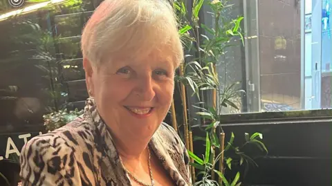 Jean Martyn A lady with short blonde hair in a leopard print blazer and gold necklace sits in a restaurant with plants in the background.