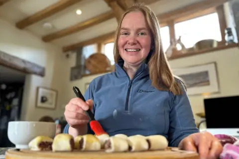 BBC A woman smiles at the camera while brushing sausage rolls