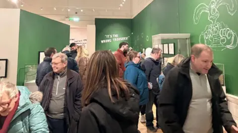 A group of museum goers shuffle past an exhibition space filled with sketches and phrases from the Wallace and Gromit films