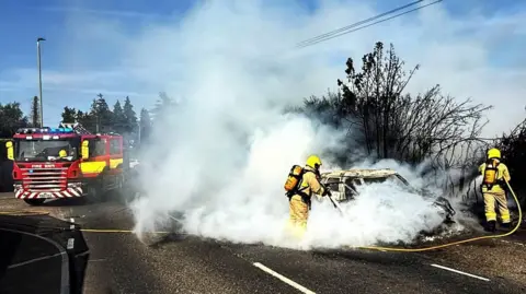 Shropshire Fire and Rescue Service Two firefighters with yellow helmets and hoses are spraying a charred car which is engulfed in smoke. It is parked at the side of a road with a blackened hedge behind it. A fire engine is parked in the middle of the road in the background