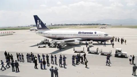 Mirrorpix via Getty Images A Britannia Airways plane stationary at an airport surrounded by security guards and baggage handlers. 