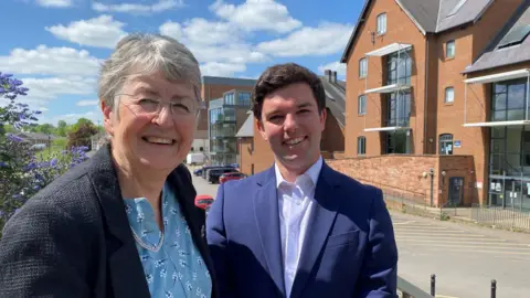 A woman with short grey hair and glasses wearing a blue patterned blouse and dark blue jacket stood next to a man with short dark hair and blue suit in front of a large brick building