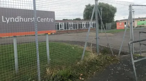 Aled Thomas The grounds of Lyndhurst Centre. There is a patch of green grass and a large sign showing 'Lyndhurst Centre'. There is a large metal fence and tarmac on the ground. 