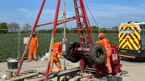 East West Rail Workers in hi-vis orange attire operate a large red drill that can be seen entering the ground. The men wear hard hats and eye protection. A field can be seen behind them as well as a van.