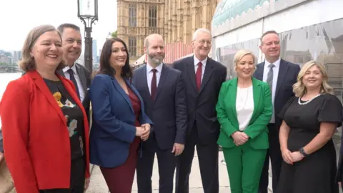 Eight people - four men and four women - post side by side for a photograph. They are standing in front of the brown Westminster building and the River Thames can be seen in the background.