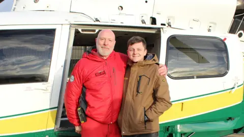 Paramedic Terry Sharpe and Jed Rayner posing for a picture in front of a GNAAS helicopter. They are holding their arms around each other and smiling. Mr Sharpe is in a red paramedic's uniform.