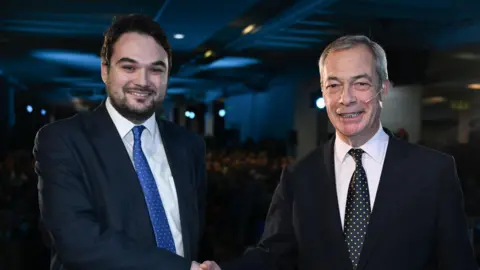 Reform UK Robin Hunter-Clarke posing for a handshake with Nigel Farage. Both are wearing dark suits. Hunter Clarke has a blue spotted tie, Farage has a black spotted tie.