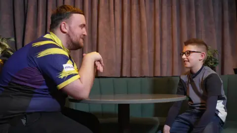 Luke Littler and Josh sat in a green cushioned booth. Luke is wearing a blue and yellow tshirt and Josh wears a grey long sleeve shirt and square framed black glasses. Both are smiling at each other