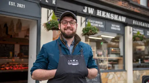 Jon Parker Lee James Henshaw wearing a dark baseball cap and butcher's apron, smiling outside his business front