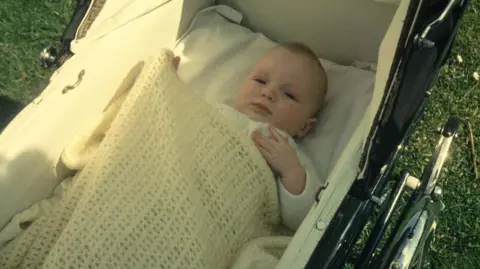 David, Jan's baby, is seen covered in a yellow blanket up to his arms, in a pram in an old colour photograph 