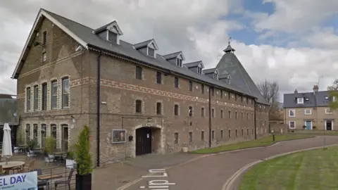 Google A long building, which is three stories, goes across the whole length of the image. The brown brick building has lots of rows of small windows and an arch entrance with a door on the left hand side. 