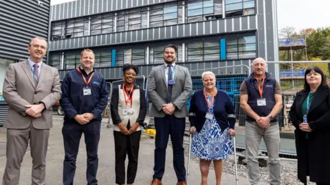 Seven people standing in front of Ercall Wood School in Wellington. Building work on an extension is going on behind them.    