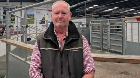 Mark Walter is standing in front of a gated enclosure at a livestock market. He is wearing a red and white checked shirt and brown waistcoat.